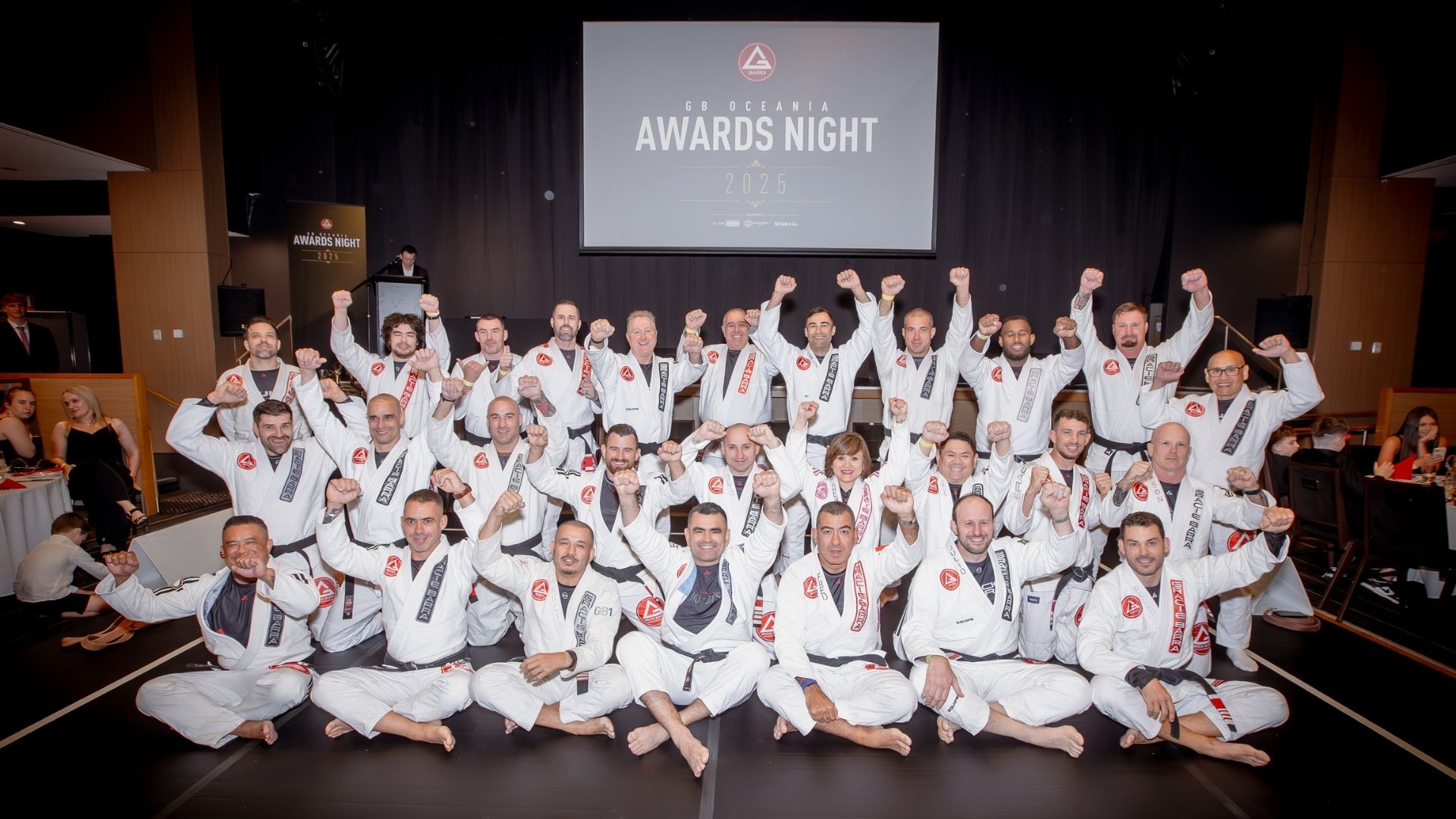 Black Belts and instructors from Gracie Barra Oceania posing together at the 2025 Awards Night, wearing white GB uniforms and raising their fists in celebration.